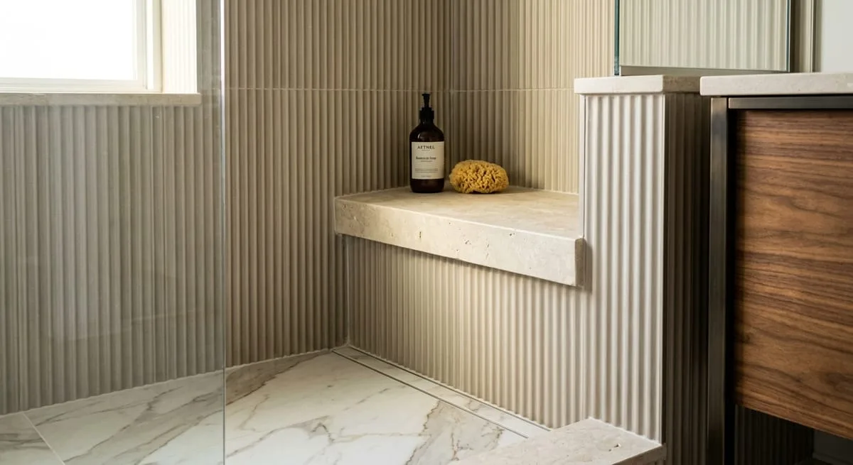 Fluted tile panels and honed stone shelf in a master bathroom shower with walnut vanity cabinet and natural stone porcelain floor